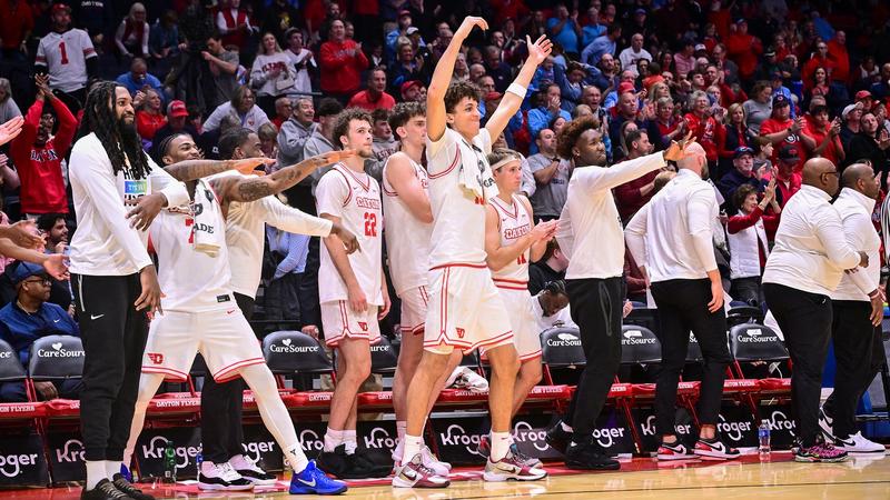Dayton men’s basketball players and coaches celebrate on the sideline with arms raised and extended as fans cheer and applaud in the stands at UD Arena.