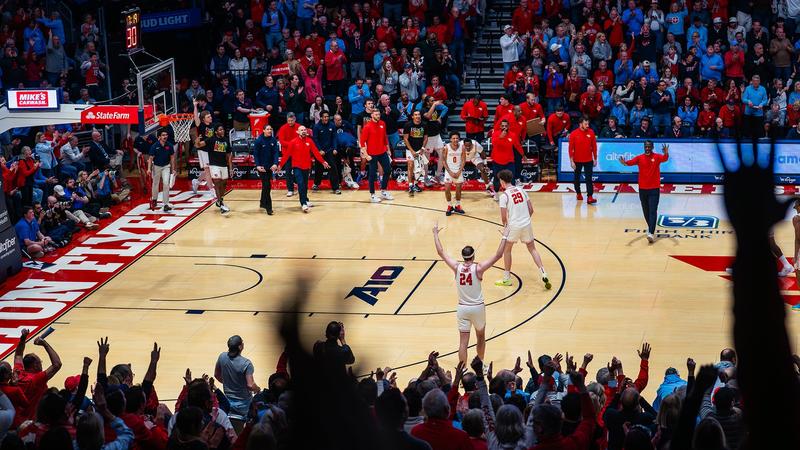 A wide-angle view of a packed basketball arena captures the home crowd celebrating as two players in white uniforms stand on the court with their arms raised toward the fans. Teammates and coaches along the bench leap to their feet in celebration, while spectators throughout the arena cheer and appl