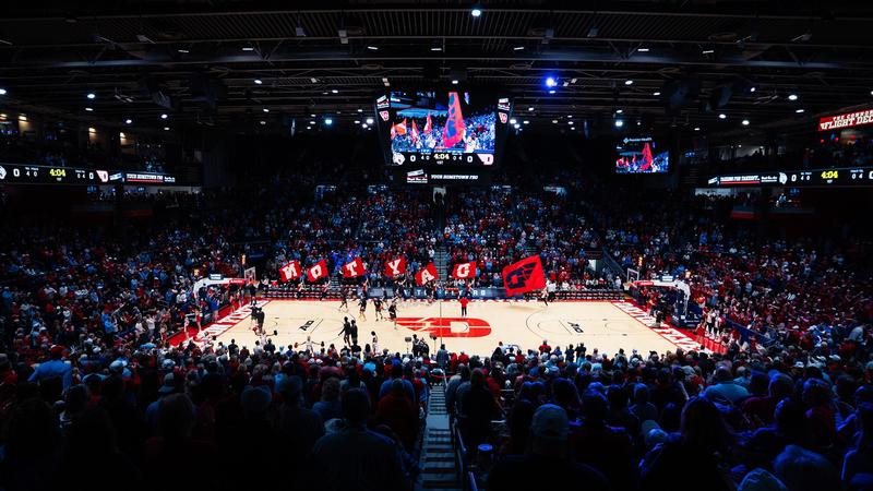 A wide-angle view of a packed basketball arena before tipoff at the University of Dayton. Fans fill the stands as members of the spirit squad run across the court waving large red flags that spell “DAYTON” alongside a Flyers logo flag. Players warm up on the court beneath a center-hung video board d