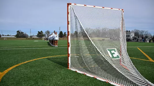 The lacrosse team stands in a single-file line facing the goal during the National Anthem