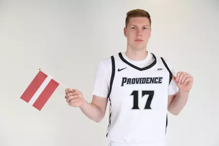 Petris Pinnis men's Basketball player holds the Latvia flag in his right hand while his other thumb is placed under his jersey to showcase it during their photoshoot.