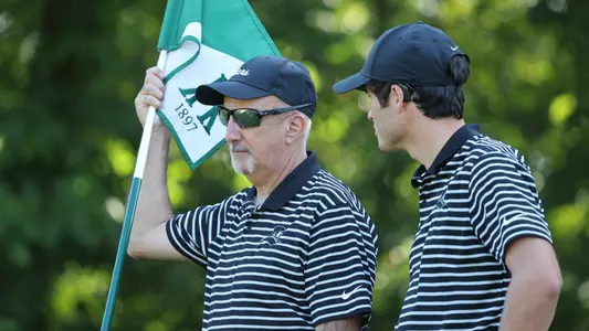 Men's golf head coach Lou Parente holding the flag stick and talking with a player
