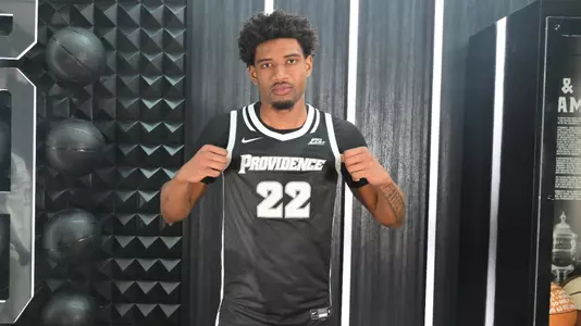 Arrinten Page poses in black providence college jersey with white trim and numbers holding the providence team name out with his hands. 