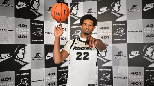 Arrinten Page poses in front of friar logo wall spinning a basketball on his finger and points at the camera in a white jersey with black trim and numbers