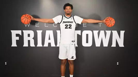 Arrinten Page poses in front of Friartown logo on the wall holding his arms out with a basketball in each hand in white jersey with black trim and numbers
