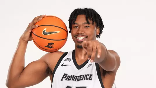 Men's basketball player Jacob Bannarbie with a basketball on his shoulder and pointing towards the camera