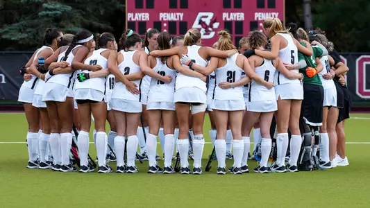 The Providence College field hockey team, dressed in white uniforms with black trim, huddles together on the field with their arms around one another.