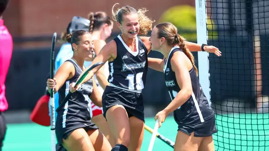 Three field hockey players dressed in black uniforms with white trim celebrate with each other after scoring a goal.