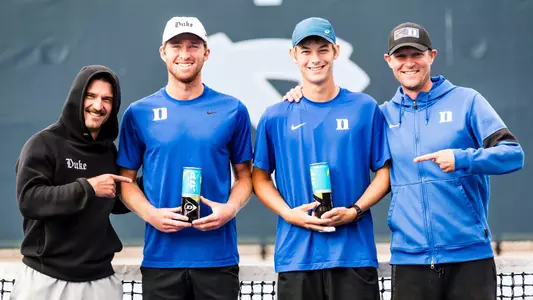 Theo Winegar and Cooper Williams pose with assistant coach TJ Pura and head coach Ramsey Smith in front of a tennis court net after capturing NCAA Doubles Championship bids.