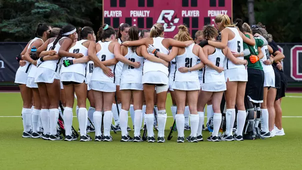 The Providence College field hockey team, dressed in white uniforms with black trim, huddles together on the field with their arms around one another.
