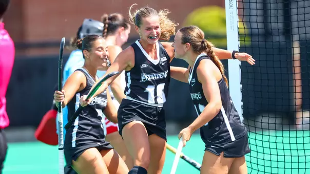 Three field hockey players dressed in black uniforms with white trim celebrate with each other after scoring a goal.