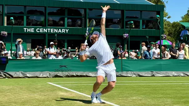 Former Duke men's tennis All-America Theo Winegar, wearing a white hat, shirt and shorts, tosses a ball up for a serve during a first round doubles match at Wimbledon.