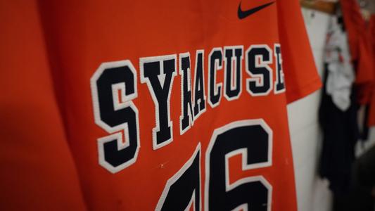An orange men's lacrosse jersey hangs in the locker room, deep in the bowels of Goldfarb Gymnasium at Johns Hopkins. Syracuse appears in bold, dark blue letters with a white stroke.