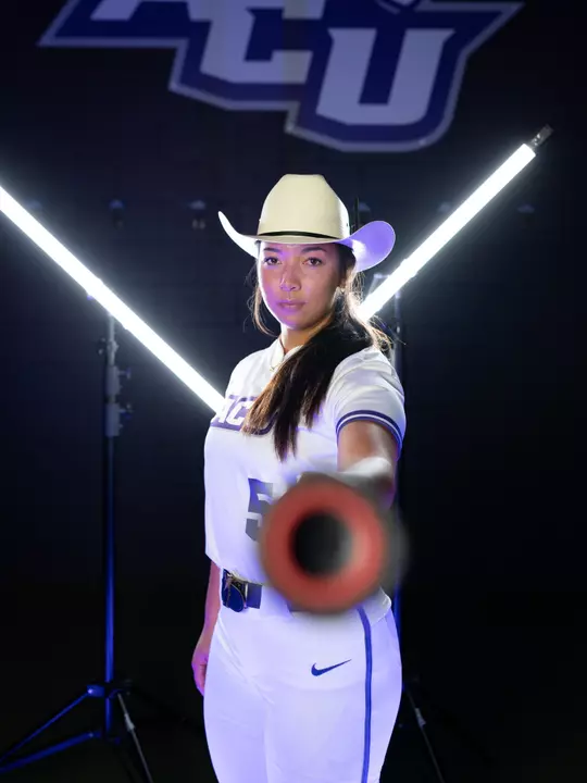 Shaylee Alani points a bat toward the camera in front of two bar lights