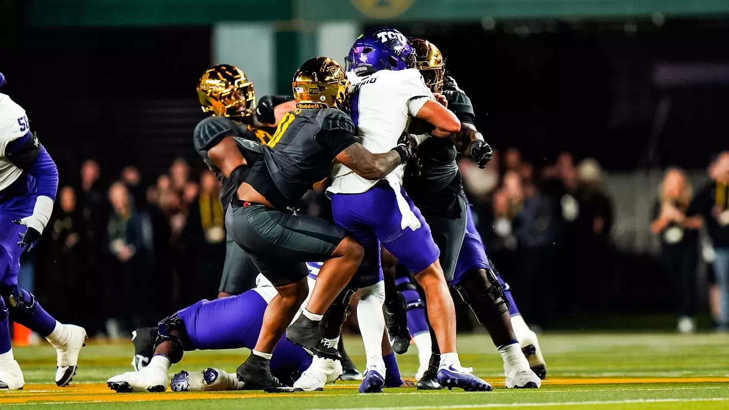 A Baylor defender in a gold helmet and dark gray uniform wraps up a TCU ball carrier in a white jersey and purple helmet during a night game. Another Baylor player closes in from behind as TCU linemen in purple pants are pushed back around them, with a blurred crowd in the stands behind the field.