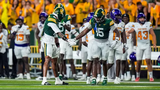 Two Baylor Bears football players in green jerseys slap hands on the field as Prairie View A&M Panthers players in white and purple stand in the background near the sideline.