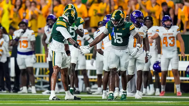 Two Baylor Bears football players in green jerseys slap hands on the field as Prairie View A&M Panthers players in white and purple stand in the background near the sideline.