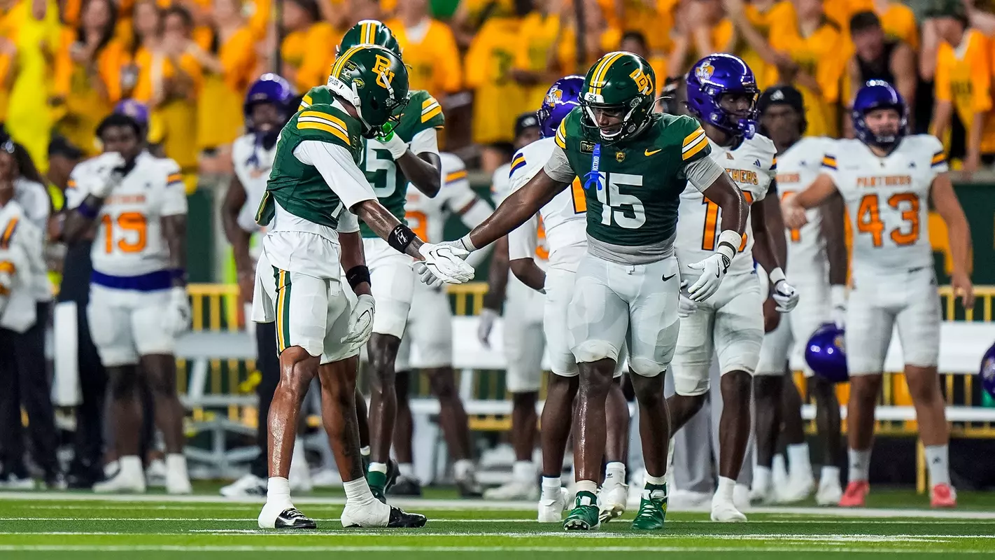 Two Baylor Bears football players in green jerseys slap hands on the field as Prairie View A&M Panthers players in white and purple stand in the background near the sideline.