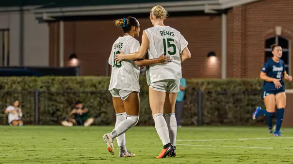 Baylor's Aryanna Jimison (No. 10) and Grace White (No. 15) walk arm in arm across the field during a night match, with the opposing team's colors visible on a nearby player in the background.