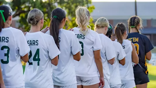 Soccer players in white 'Bears' jerseys stand arm-in-arm in a line facing away from the camera, next to a player in a dark 'Sic 'Em' jersey, before a match on a grass field with trees and a building in the background