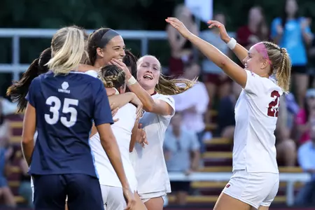 A group of players celebrates a goal, with goal scorer Claire Mensi smiling among the crowd.