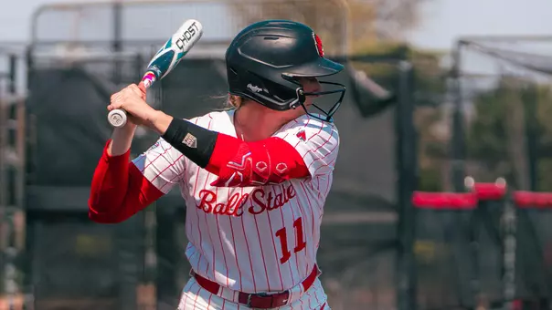 Ella Whitney batting in her white pinstripe uniform at the Ball State Softball Stadium.
