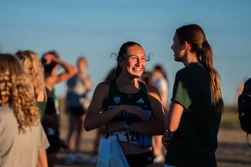 A female runner smiles and talks with another woman at an outdoor track event surrounded by other athletes and spectators.