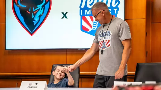 Coach Holtmann (standing on the right) fistbumps Frankie (sitting on the left) in front of a "Team IMPACT" backdrop. Frankie has a smile on his face with a name card on the table in front of him