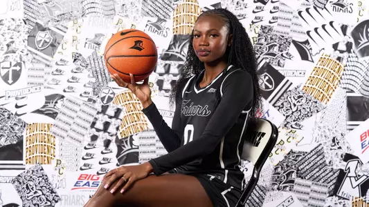 Sabou Gueye poses in front of a Friar-branded backdrop, seated in a chair holding a basketball with a Friar head logo on it.
