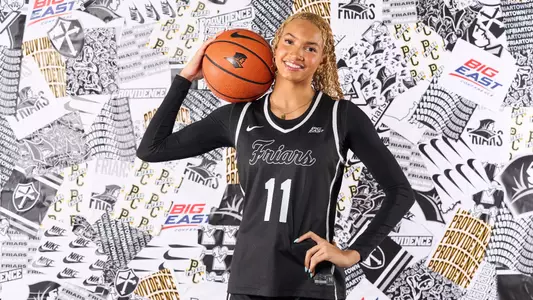 Riley Hamburger posing in front of a Providence College athletics-branded backdrop in her black uniform with the number 11 on it, holding a basketball on her right shoulder.