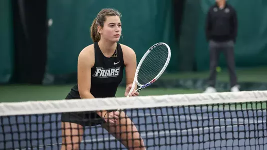 Tennis player Camila Barrera waits in an athletic stance behind the net for her opponent to serve during an indoor match.
