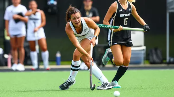 Martu Torre Festa, dressed in a white uniform with black trim, passes a field hockey ball with her stick.