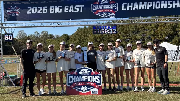 A group photo of the 2026 Women's Cross Country BIG EAST Champions holding the trophy in front of the finish line.
