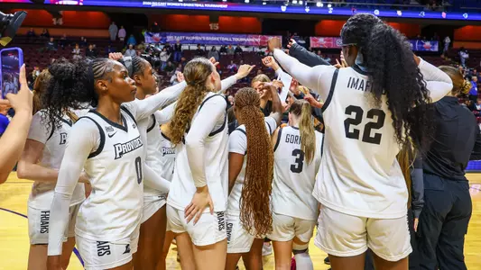 The women's basketball team dressed in white uniforms huddles together for a pregame cheer before playing a game.