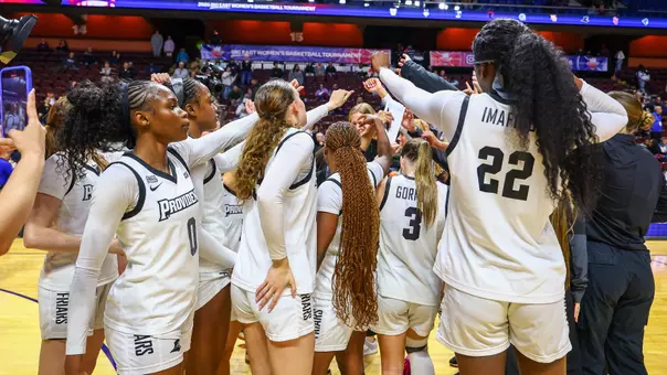 The women's basketball team dressed in white uniforms huddles together for a pregame cheer before playing a game.