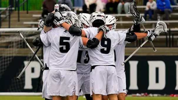 Lacrosse team celebrates a goal versus Denver in a huddle. Rhett Chambers and Richie Joseph are in the front of the picture with their backs turned. the team is wearing white uniforms with white helmets on chapey field at anderson stadium