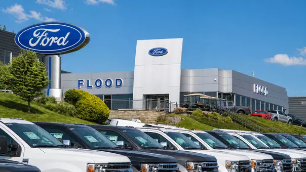 Flood Auto Group Picture for Release exterior shot of Flood Ford with signage with both "Flood" and "Ford" lettering feature white scrip for Ford and blue capitals letters for Flood. a row of pickup trucks is in the foreground with a few more in front of the right side of the building