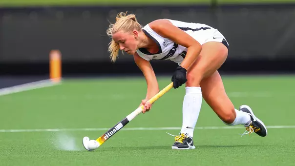 Saar Koper, wearing a white uniform with black lettering and trim, sweeps the ball with her field hockey stick.