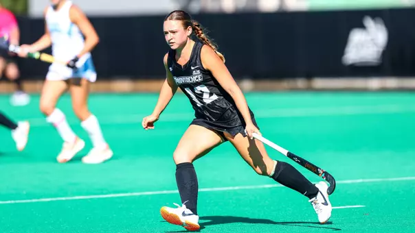 Carly van Benten, dressed in a black uniform with white trim, runs on an astroturf field with her field hockey stick low to the ground while playing defense.