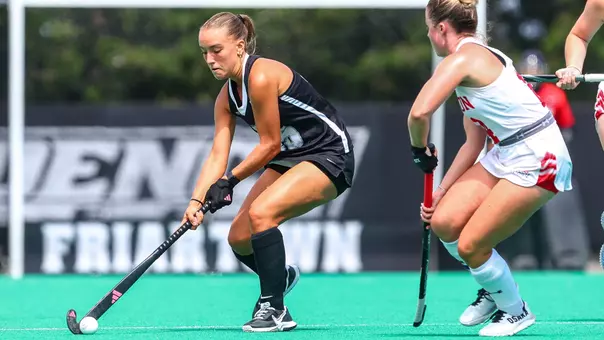 Field hockey player Riley Givens, dressed in a black uniform with white trim, dribbles the ball with her stick while being defended by a player from Boston University who is dressed in a white uniform with red trim.