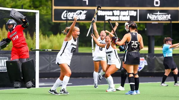 Three Providence College field hockey players dressed in white uniforms with black trim raise their arms in celebration after scoring a goal.