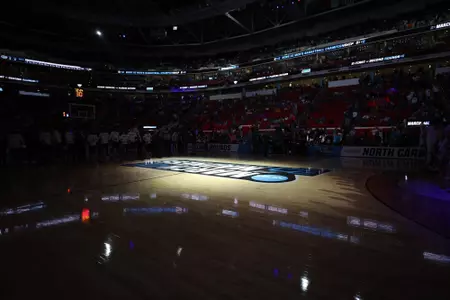 A spotlight shines on the March Madness center court logo inside a dark Lenovo Center in Raleigh before Florida's 2025 NCAA Tournament game vs. UConn.