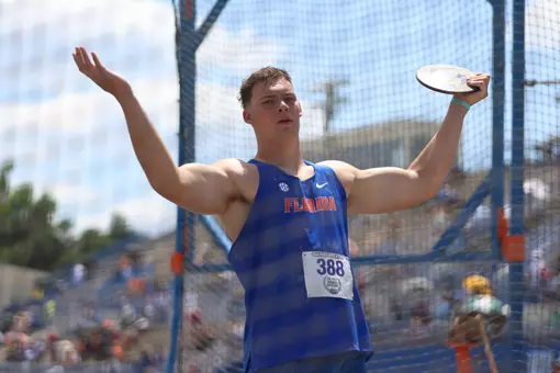 A Florida Gators men's discus thrower in blue holds his hands above his head with the discuss in his left hand.