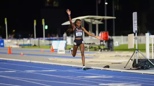 A female athlete in black uniform has arms extended as she crosses finish line on blue track at night