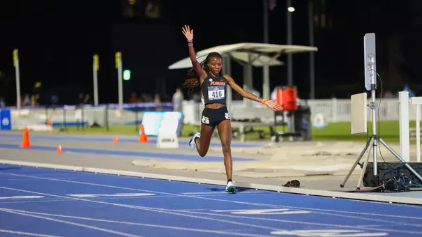 A female athlete in black uniform has arms extended as she crosses finish line on blue track at night