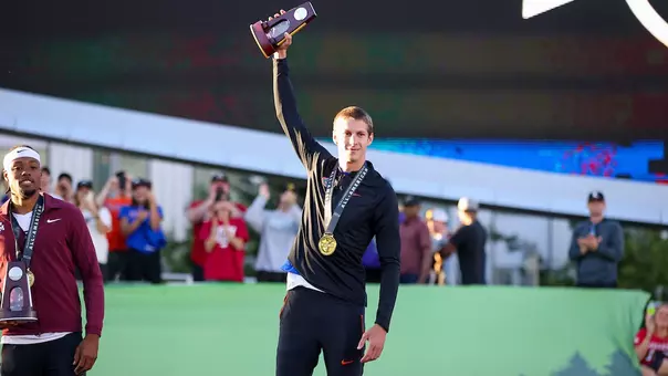 A male athlete in a black Florida Gators track suit holds a trophy above his head in the sunlight