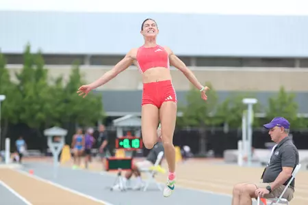 A female athlete in a red uniform mid-jump over a sand pit