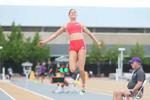 A female athlete in a red uniform mid-jump over a sand pit