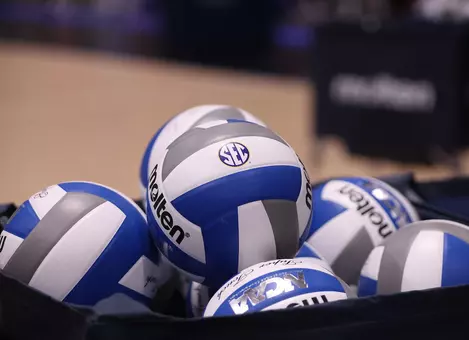 A blue and white volleyball with a conference logo in the center of a basket of volleyballs.