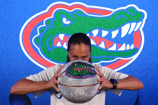 Head Women's Basketball Coach Tammi Reiss poses in front of the Gators' logo holding a diamond basketball